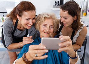 Resident using a mobile device with support from others, representing salon and personal care services at Moose Lake Village.